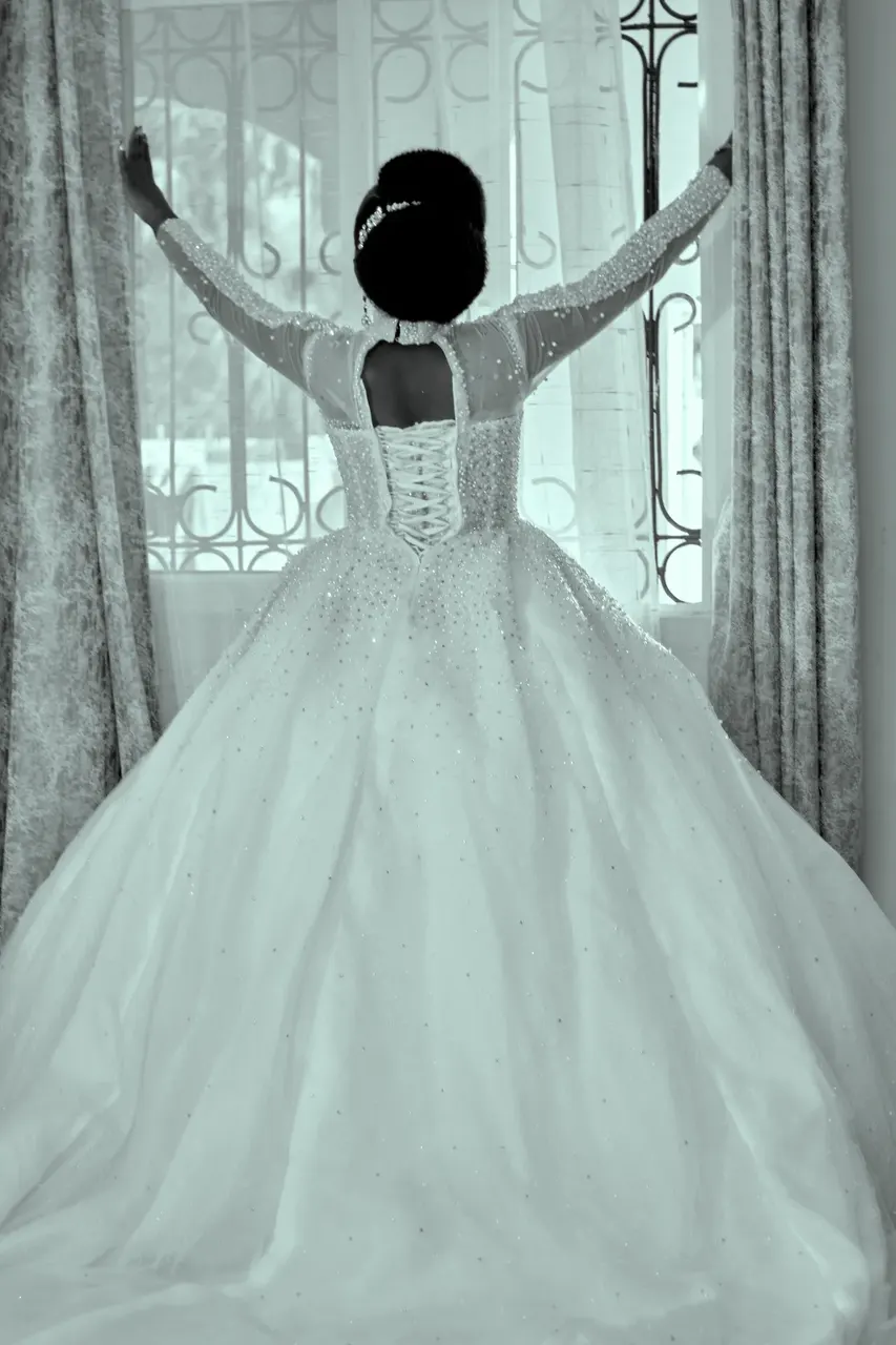 Bride with arms raised by the window, back of her lace wedding gown in black and white